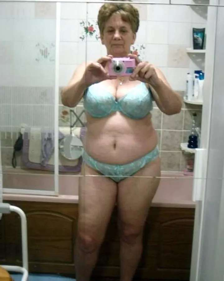 Femme âgée, en sous-vêtements bleu clair, tenant un appareil photo rose, prenant un selfie dans une salle de bain.