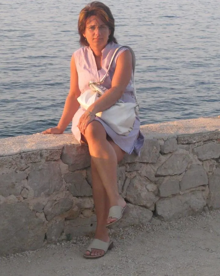 Femme aux cheveux bruns, portant des lunettes, assise sur un mur de pierre près de l’eau, vêtue d’une robe lilas.