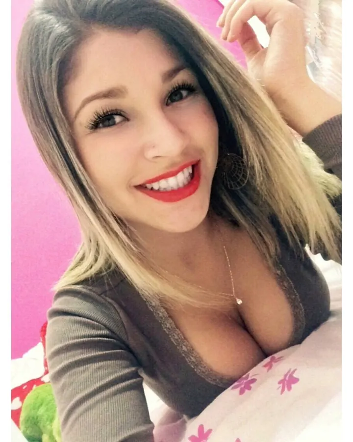 Femme aux cheveux blonds ombrés, sourire éclatant, vêtue d'un haut marron profond, pose devant un fond rose vif.