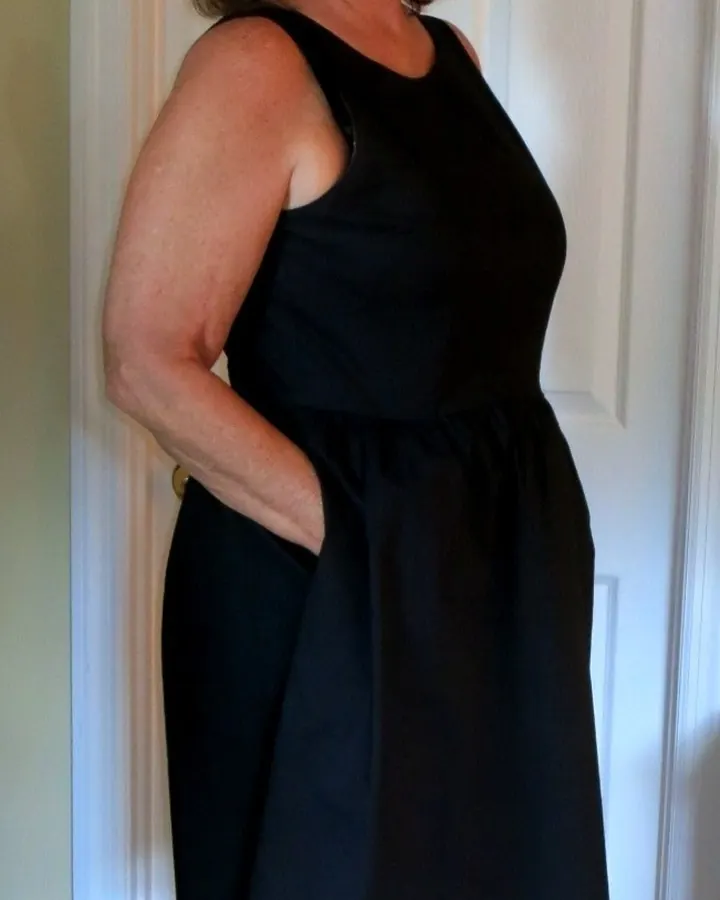 Femme en robe noire, lunettes, souriant, debout devant une porte blanche.