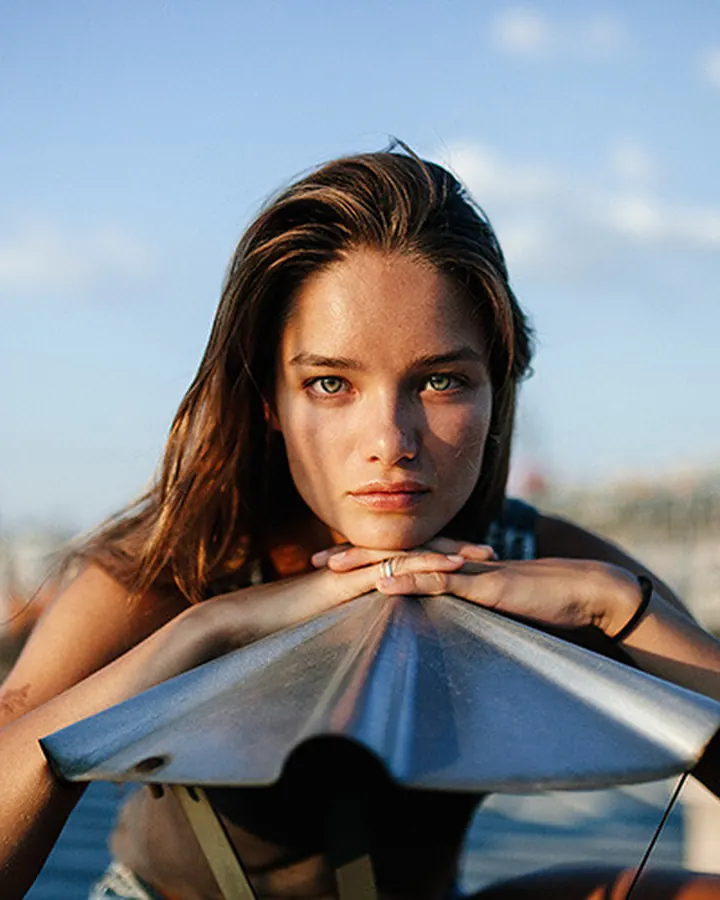 Jeune femme aux yeux bleus, en maillot noir, posant près de l'eau, regard fixe vers l'horizon.