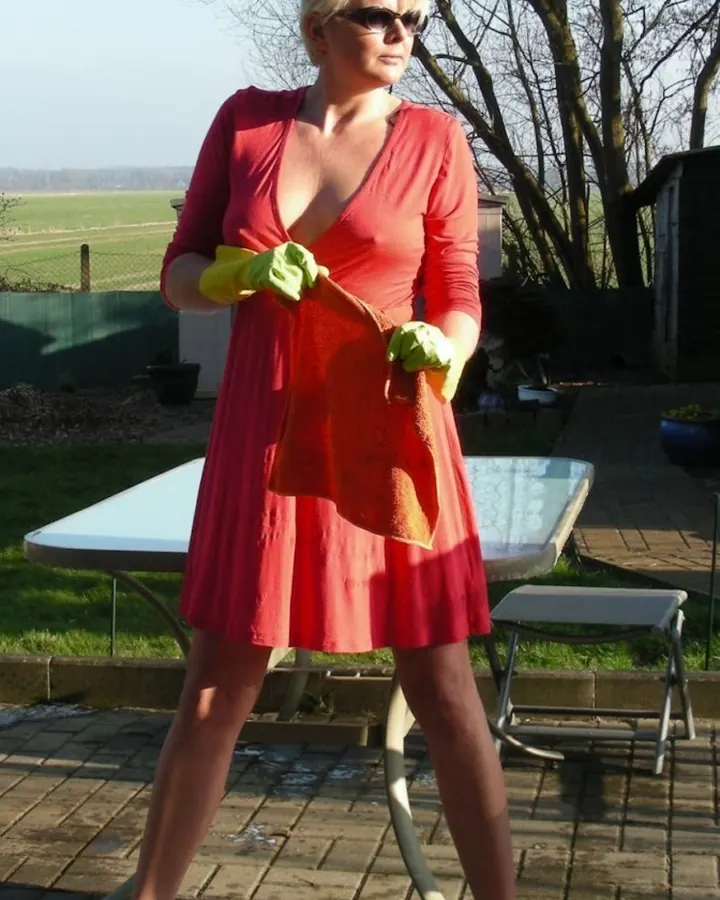 Femme blonde aux cheveux platine, en robe rouge profondément décolletée, portant des gants jaunes, en train de nettoyer une table en extérieur.