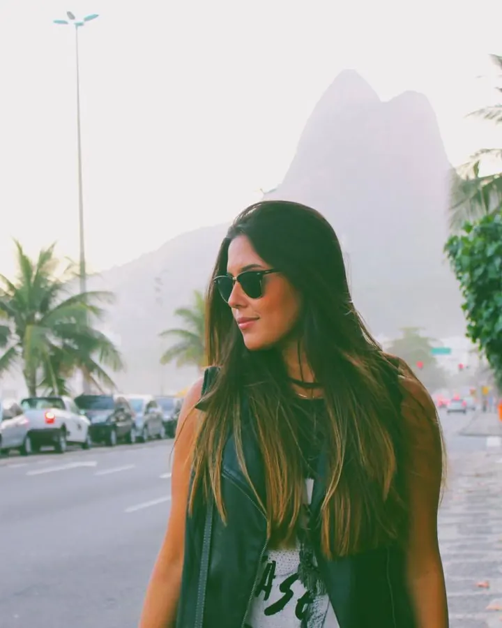 Femme latine aux cheveux châtains, en veste en cuir noir, souriante, sur une rue bordée de palmiers avec des montagnes en arrière-plan.