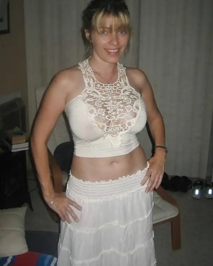 Jeune femme blonde aux cheveux longs, en tenue blanche avec un haut crop-top et une jupe courte, portant des collants à mailles, debout dans une forêt.