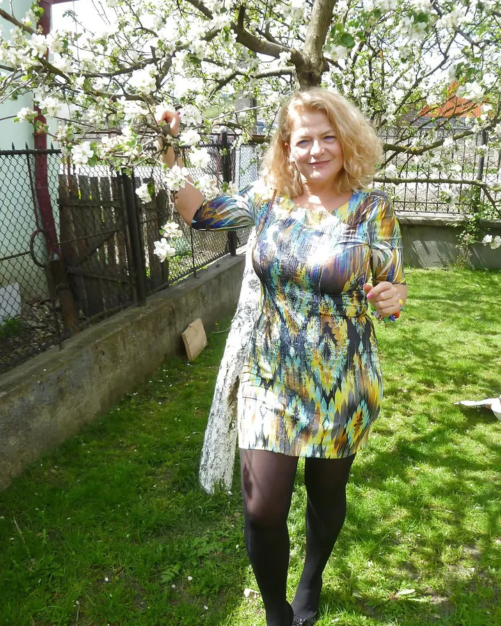Femme blonde en robe colorée, debout sous un arbre en fleurs, tenant une branche blanche.