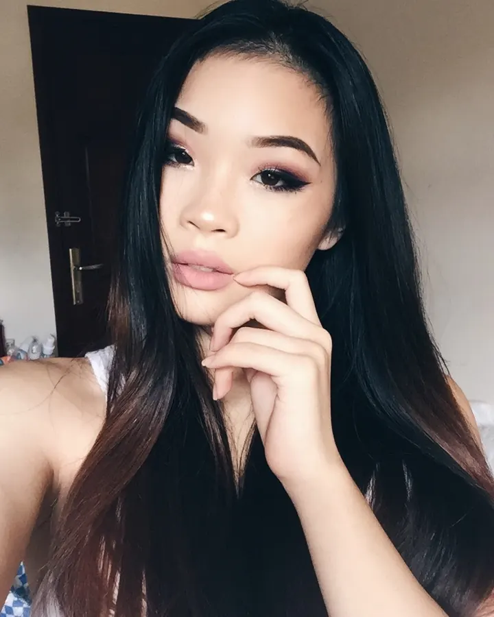 Jeune femme asiatique aux cheveux longs noirs, maquillage prononcé, prenant un selfie avec une main sur la bouche.