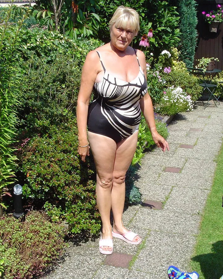 Femme blonde aux cheveux courts, portant un maillot de bain noir et blanc à rayures, debout dans un jardin fleuri.