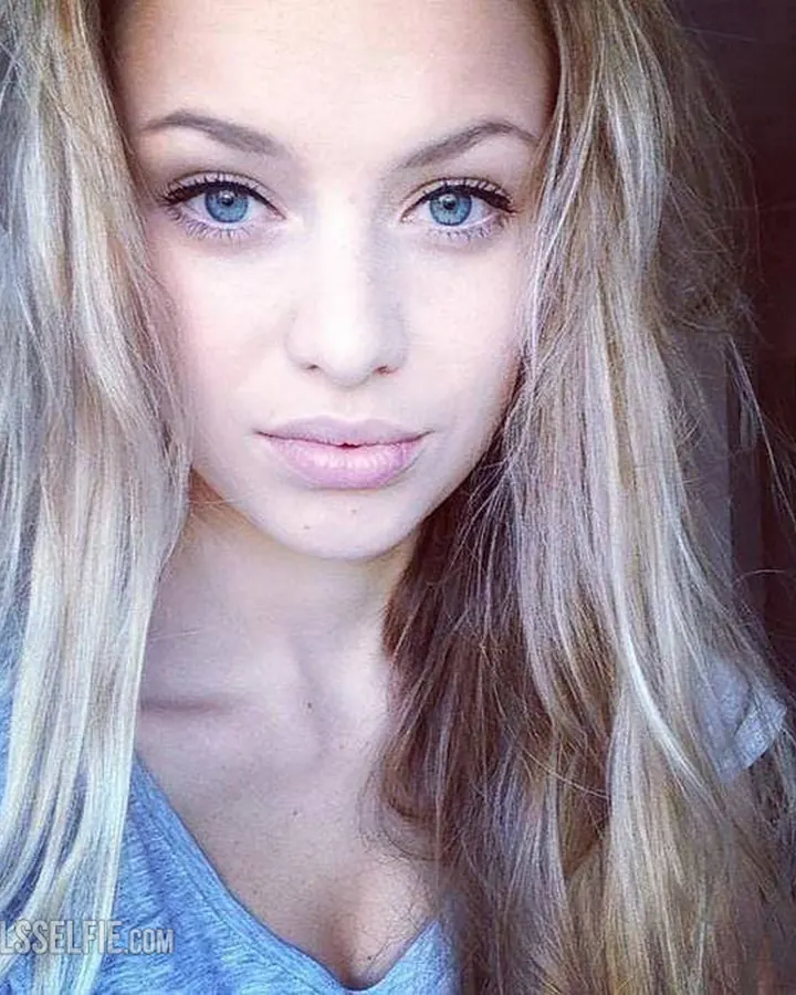 Portrait proche d'une jeune femme aux cheveux blonds et yeux bleus, portant un haut bleu clair, regardant directement l'objectif.