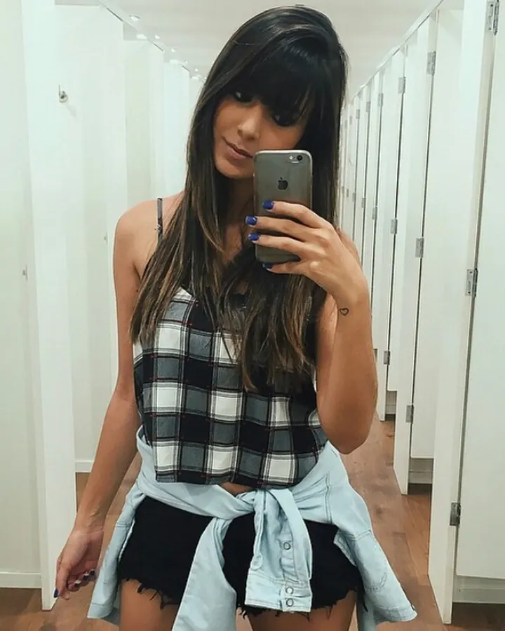 Femme aux cheveux longs bruns et frange, portant un haut à carreaux noir et blanc noué à la taille, prenant un selfie devant un miroir dans un couloir de vestiaires.