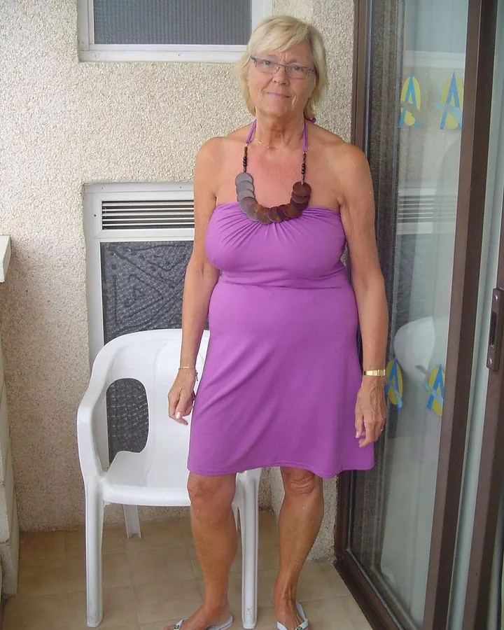 Femme aux cheveux blancs courts, portant un haut violet et une jupe assortie, debout sur une terrasse en regardant l'objectif.