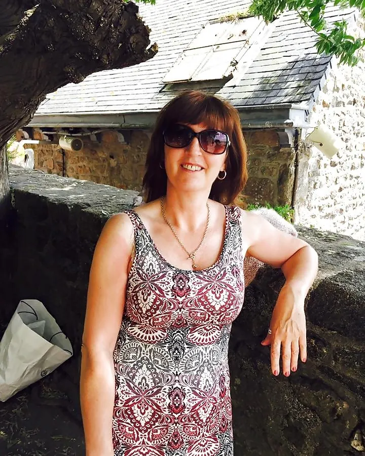 Femme aux cheveux châtains, en robe à motifs, lunettes de soleil, souriant, appuyée sur un mur de pierre devant une maison ancienne.