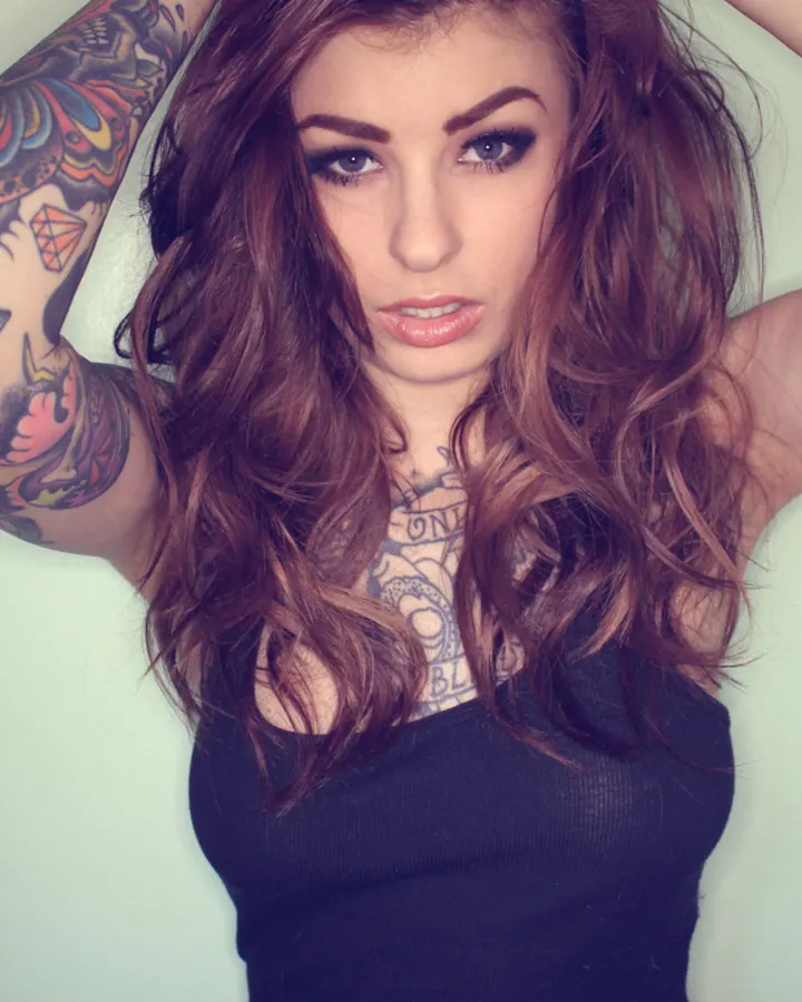 Femme aux cheveux châtains ondulés, regard intense, tatouages colorés sur le bras, portant un débardeur noir, mains dans les cheveux.