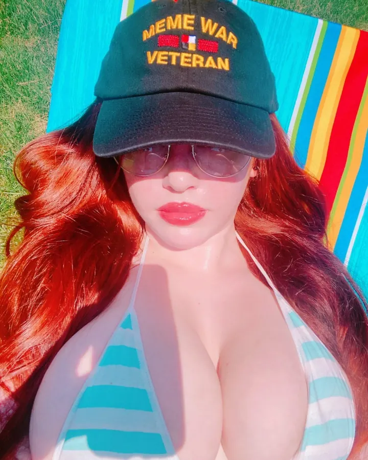 Femme aux cheveux roux longs, en maillot de bain turquoise et blanc, casquette verte "MEME WAR VETERAN", lunettes de soleil, allongée sur une serviette multicolore.