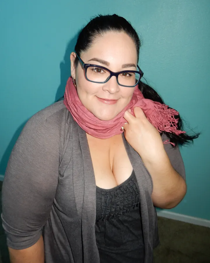 Femme aux cheveux noirs, portant des lunettes et un foulard rose, souriante devant un mur bleu.