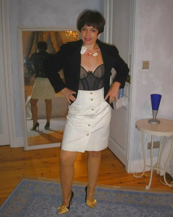 Femme aux cheveux courts, en veste noire, corset noir et jupe blanche, posant devant un miroir dans une chambre.