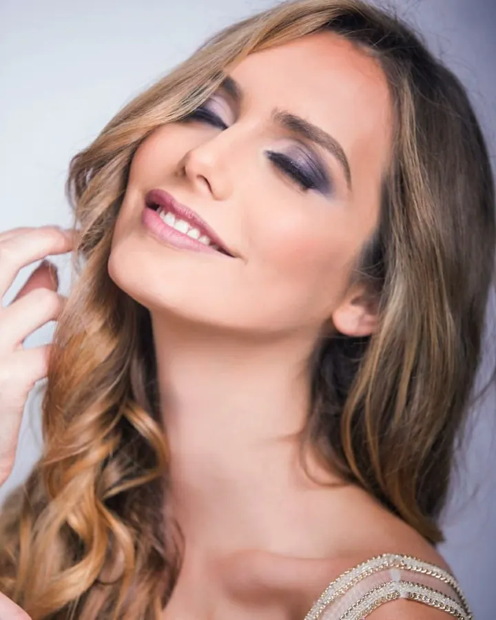 Femme aux cheveux blonds, maquillage prononcé, souriante, en haut orné, posant près d'une fenêtre.