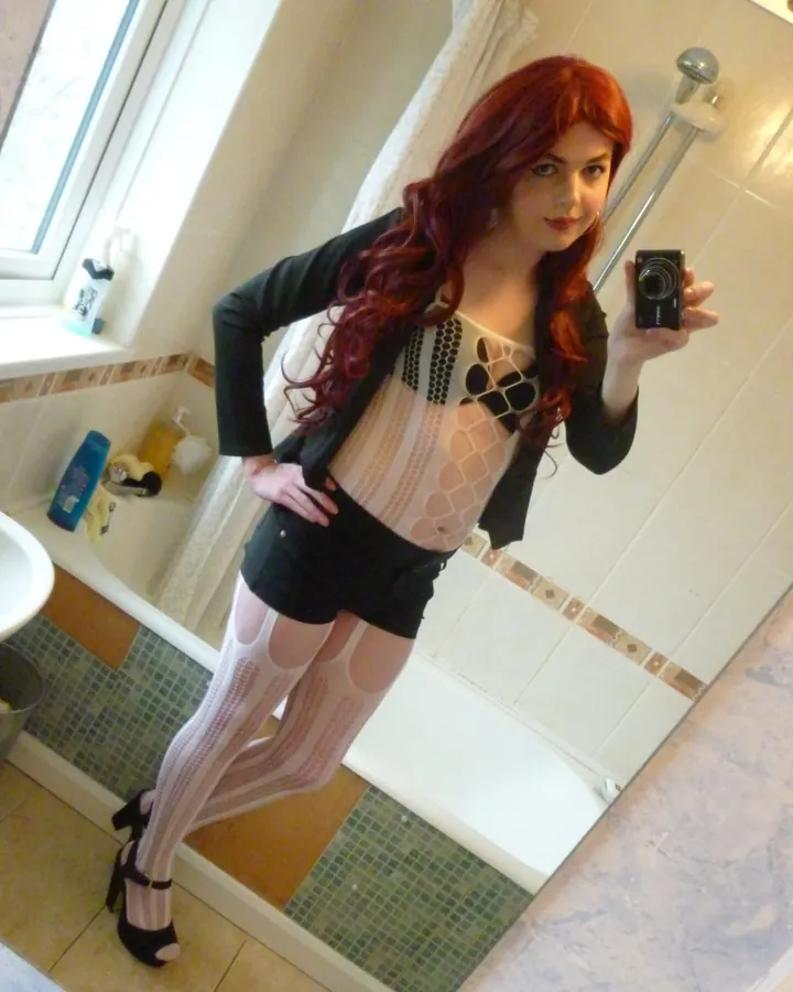 Femme blonde aux cheveux roux, souriante, robe noire, collants résille, talons hauts, assise sur canapé rouge, coussins décoratifs.