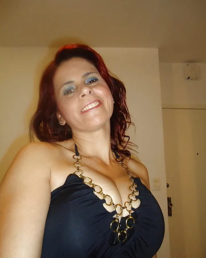 Femme aux cheveux roux, souriante, robe noire, collier chaîne dorée, intérieur simple, lumière douce.
