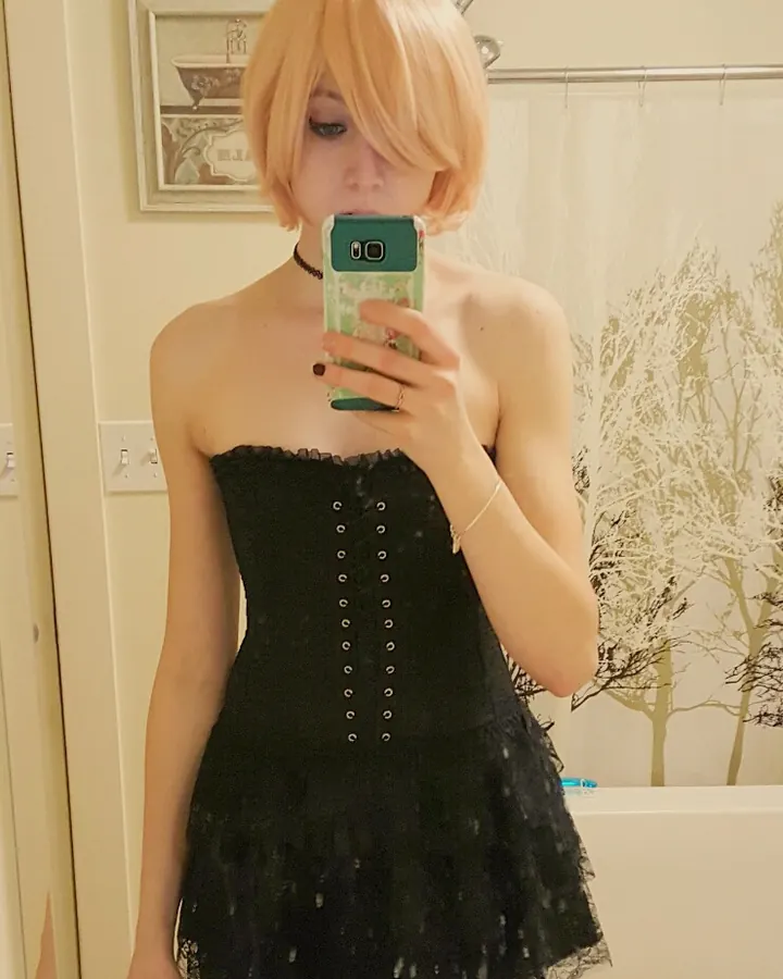 Jeune femme aux cheveux blonds courts prenant un selfie dans une salle de bain, vêtue d'une robe noire à lacets.