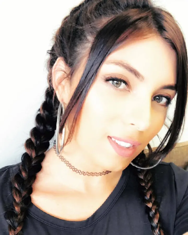 Femme aux cheveux bruns, en haut noir, avec des tresses, un collier fin et des boucles d'oreilles grandes, regardant l'appareil, souriante.
