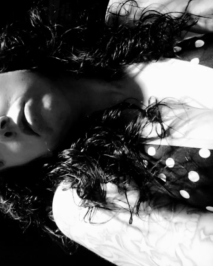 Photo noir et blanc d'une femme aux cheveux bouclés, allongée, portant un haut à pois, le regard fixe, un tatouage visible sur le bras.
