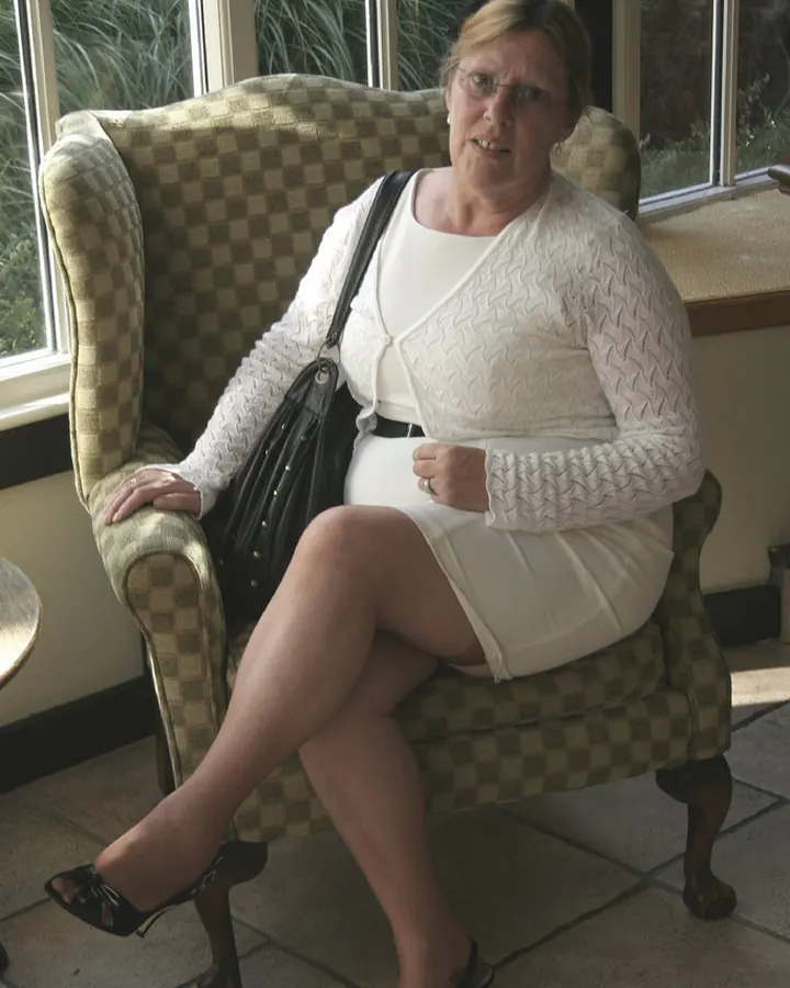 Femme blonde en tenue blanche, assise sur une chaise à carreaux, portant des lunettes, dans un cadre extérieur.
