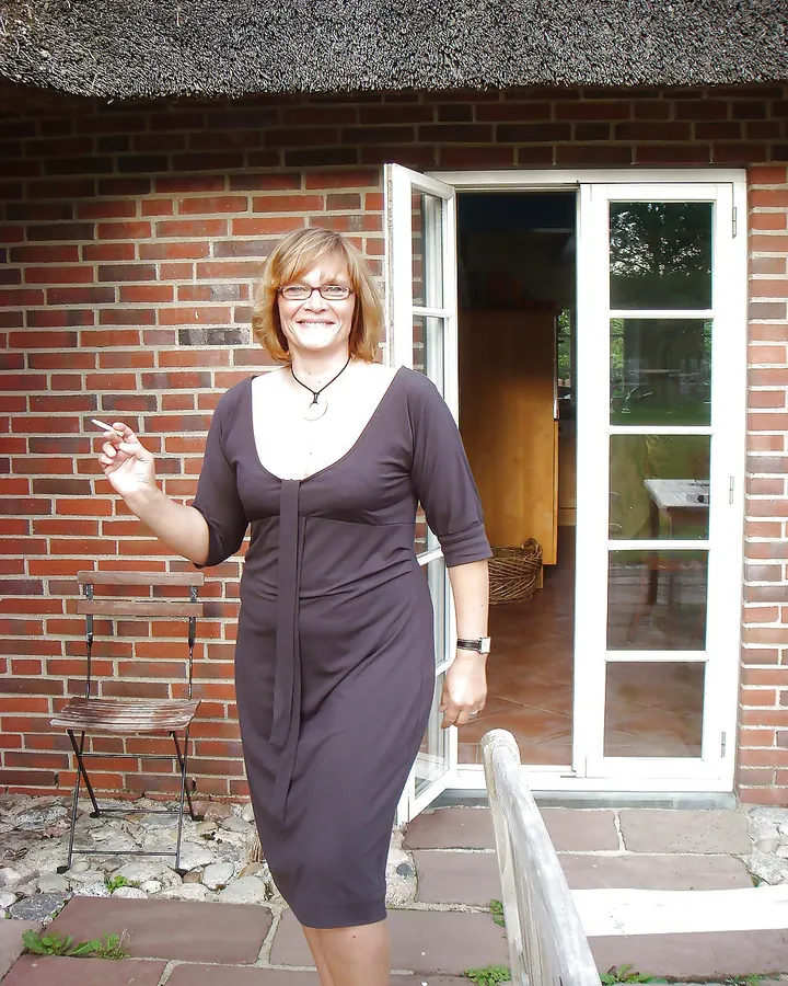 Femme de face, cheveux châtains courts, souriante, tenant une cigarette, devant une porte vitrée d’une maison en briques.