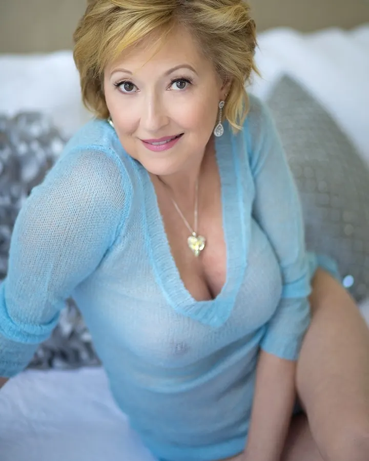 Femme aux cheveux blonds courts, en pull bleu clair transparent, assise sur un lit, regardant l’objectif avec un sourire tendre.