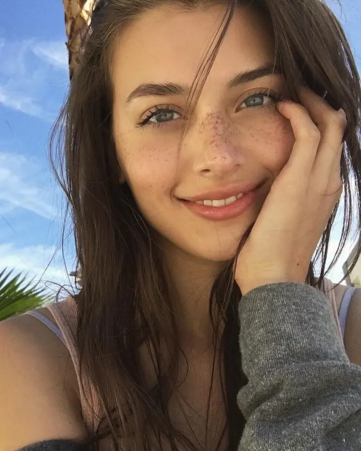 Jeune femme aux yeux bleus et cheveux châtains, souriante, tenant son visage avec la main, sous un ciel bleu.