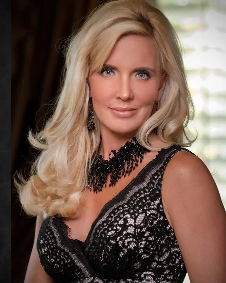 Femme blonde aux yeux bleus, vêtue d'une robe noire à dentelle, souriante, posant devant un fond sombre avec lumière douce.