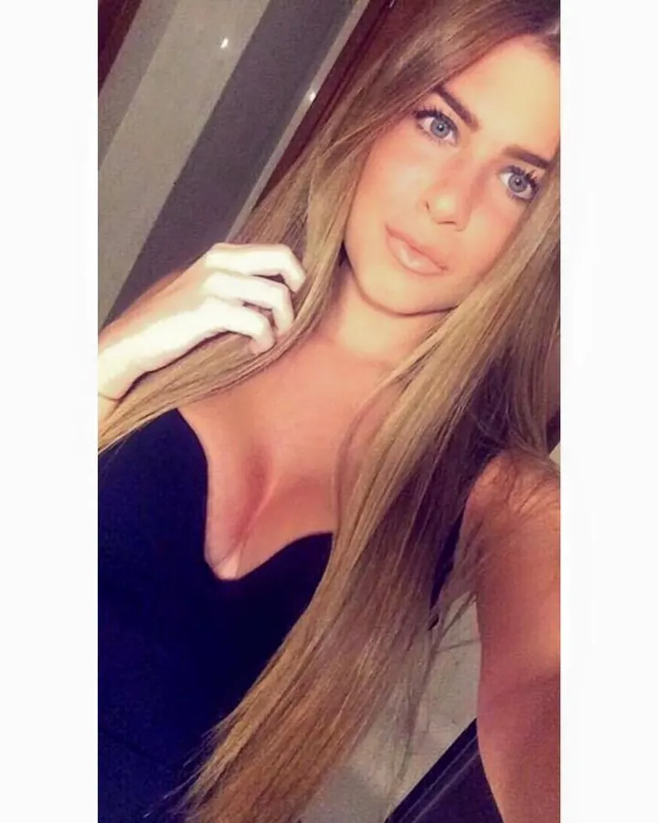 Femme aux cheveux blonds, souriante, vêtue d'un haut noir, posant dans un cadre intime.