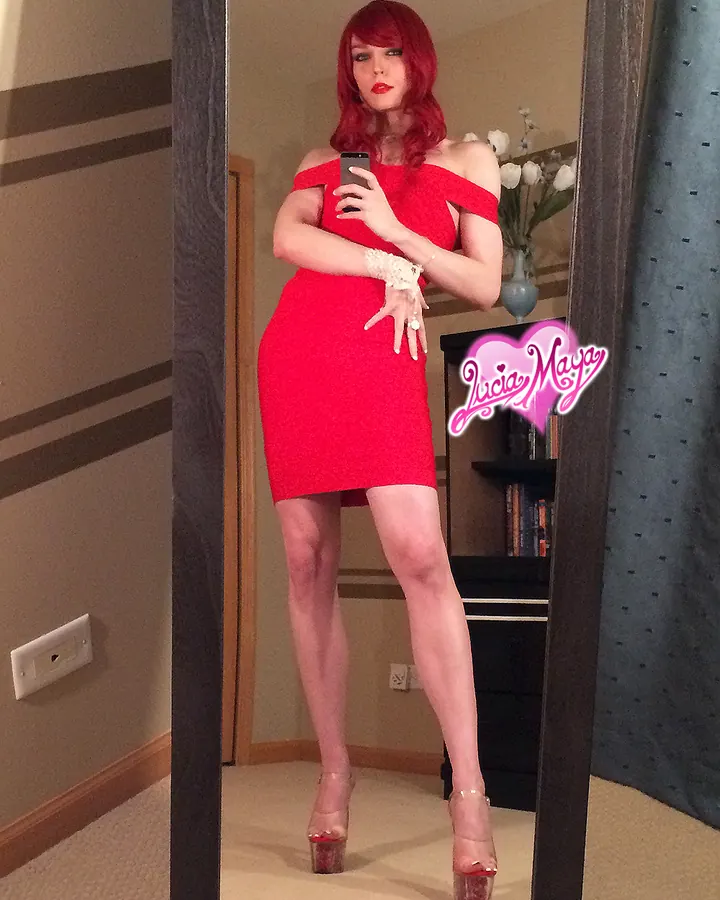 Femme aux cheveux roux portant une robe rouge, prenant un selfie devant un miroir dans une pièce aux murs beiges.