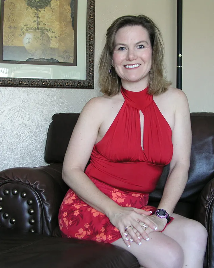 Femme souriante aux cheveux bruns, portant une robe rouge à col halter, assise sur un canapé en cuir noir dans un salon.