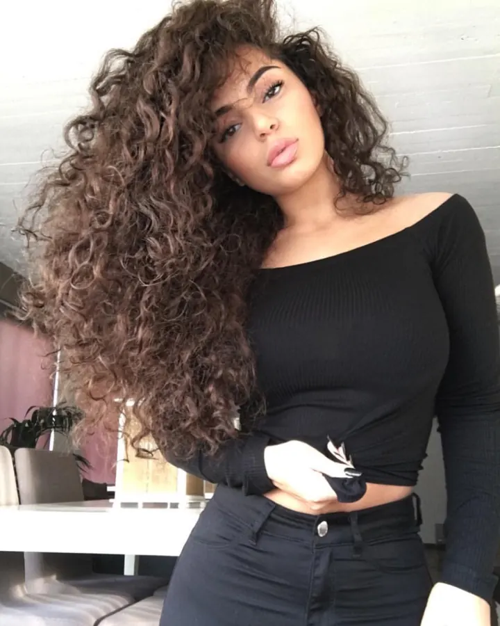 Femme aux cheveux bouclés, top noir, pantalon noir, main sur la taille, regard intense