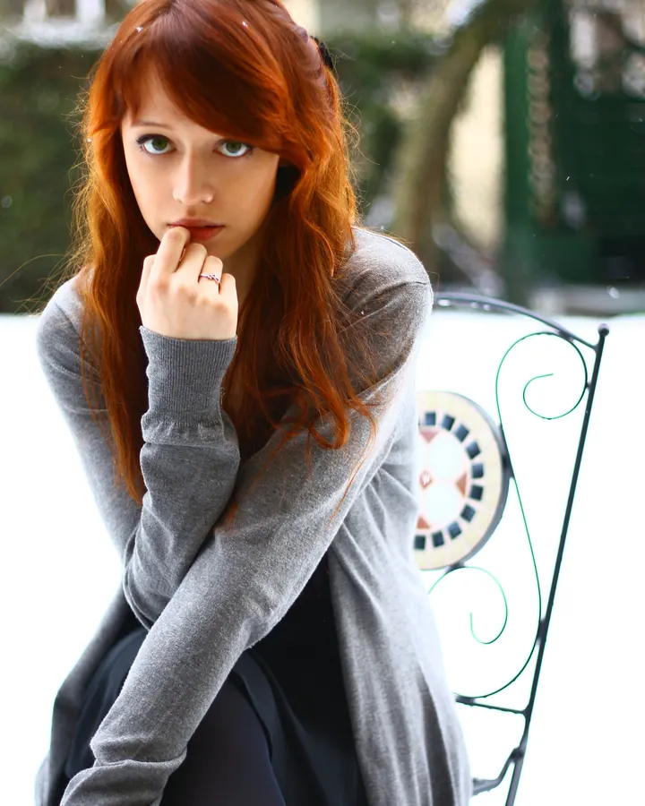 Femme aux cheveux roux, en pull gris, assise sur une chaise métallique dans la neige, main sur la bouche