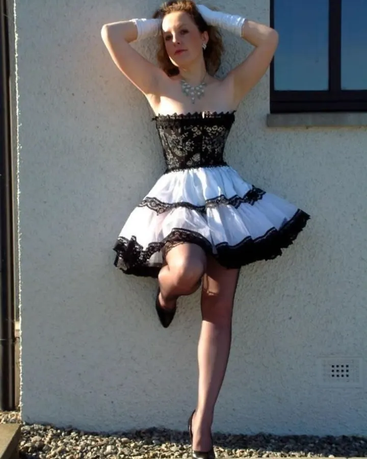 Femme en robe noire et blanche, les mains derrière la tête, posant contre un mur gris