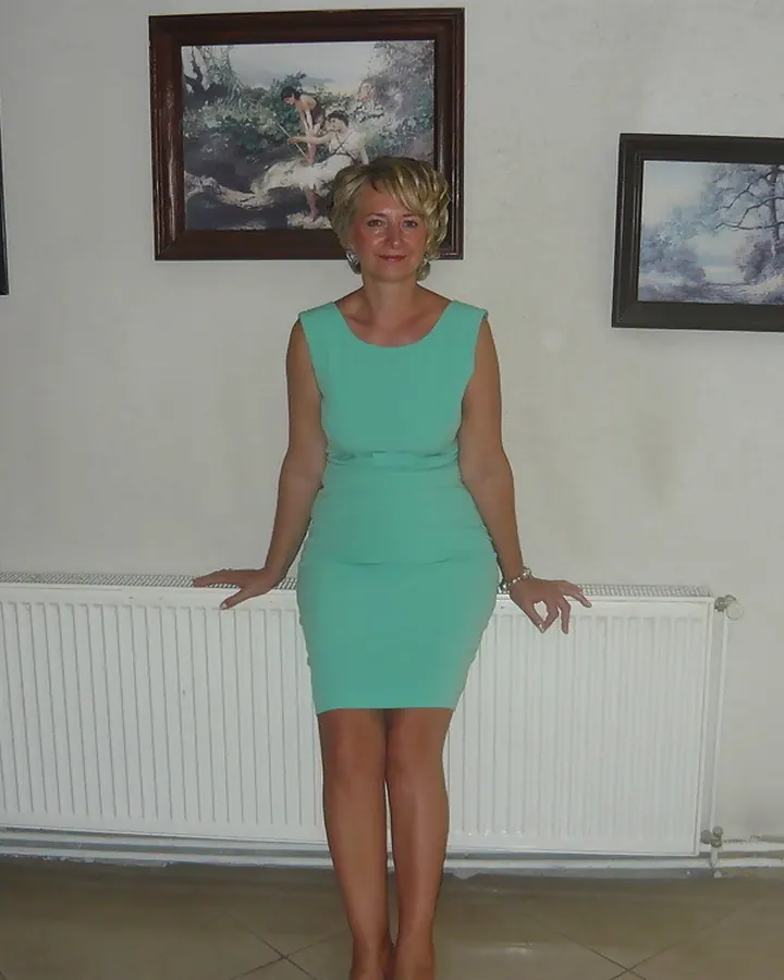 Femme souriante en robe turquoise, debout devant un radiateur blanc avec des tableaux au mur