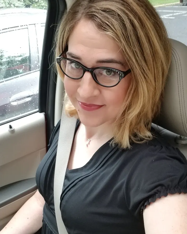 Femme aux cheveux blonds, vêtue d'un haut noir, prenant un selfie dans un miroir, portant un collier doré.