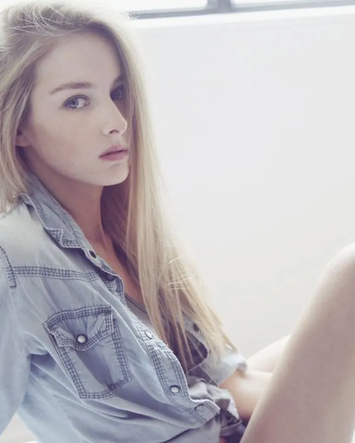 Jeune femme blonde aux yeux bleus, vêtue d'une chemise en jean, assise sur un lit blanc dans une pièce lumineuse.