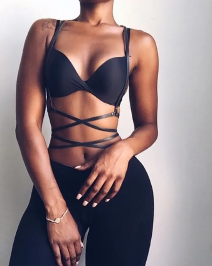 Femme africaine aux cheveux bruns-noirs, vêtue d'un soutien-gorge noir et de leggings, tenant sa main sur son ventre, devant un mur blanc.
