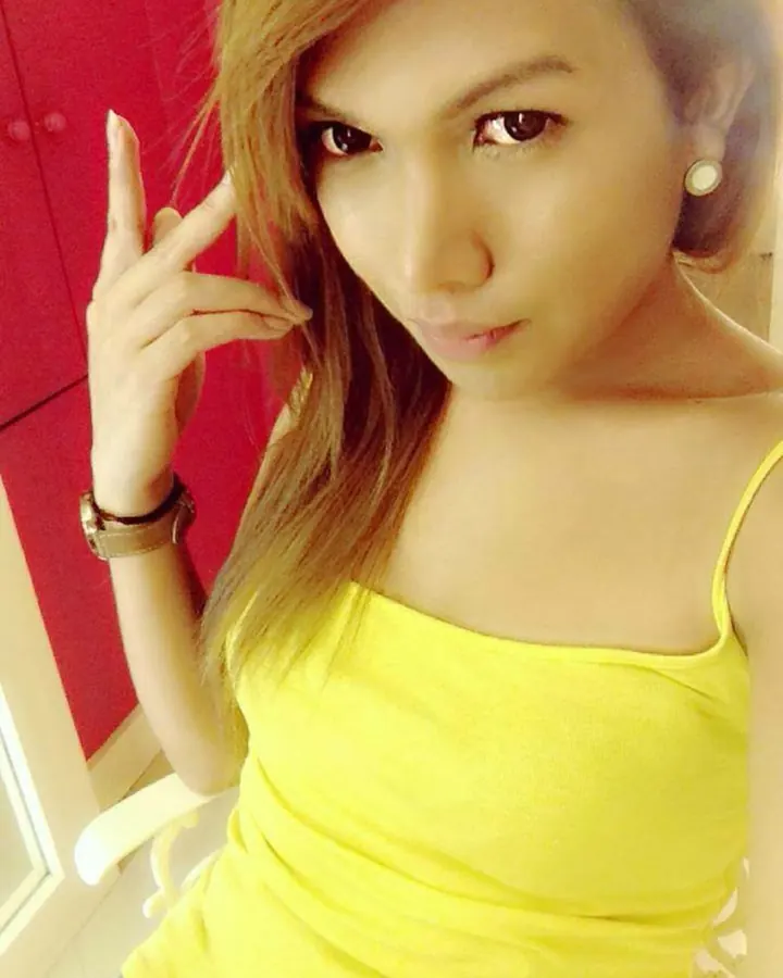 Jeune femme latino en haut jaune, cheveux châtains, regard direct, faisant un signe de la main devant un fond rouge.