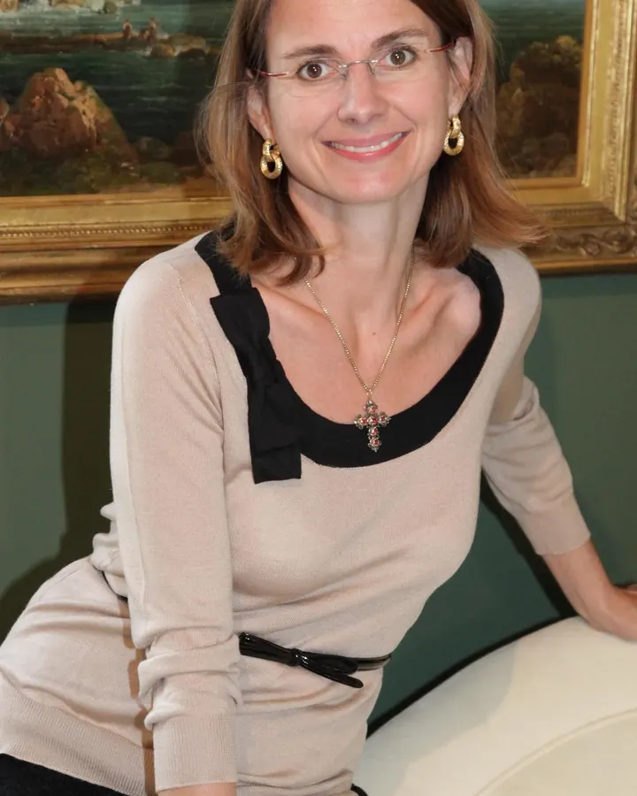 Femme aux cheveux châtains, vêtue d'un haut beige et d'une jupe noire, souriant devant un tableau paysager.