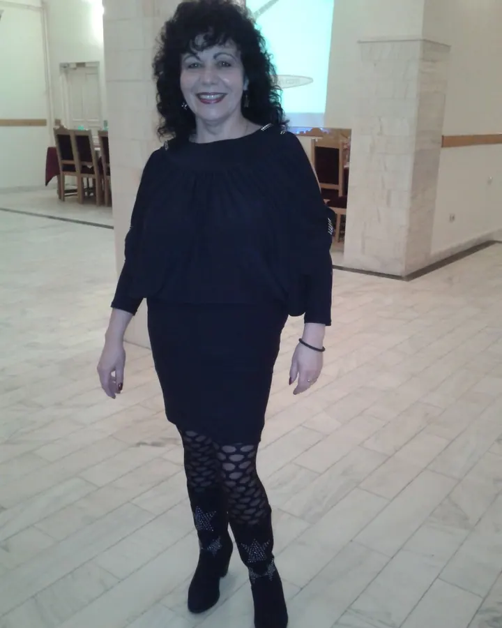 Femme aux cheveux noirs bouclés portant une robe noire, collants à motifs et bottes, souriante dans une salle intérieure.