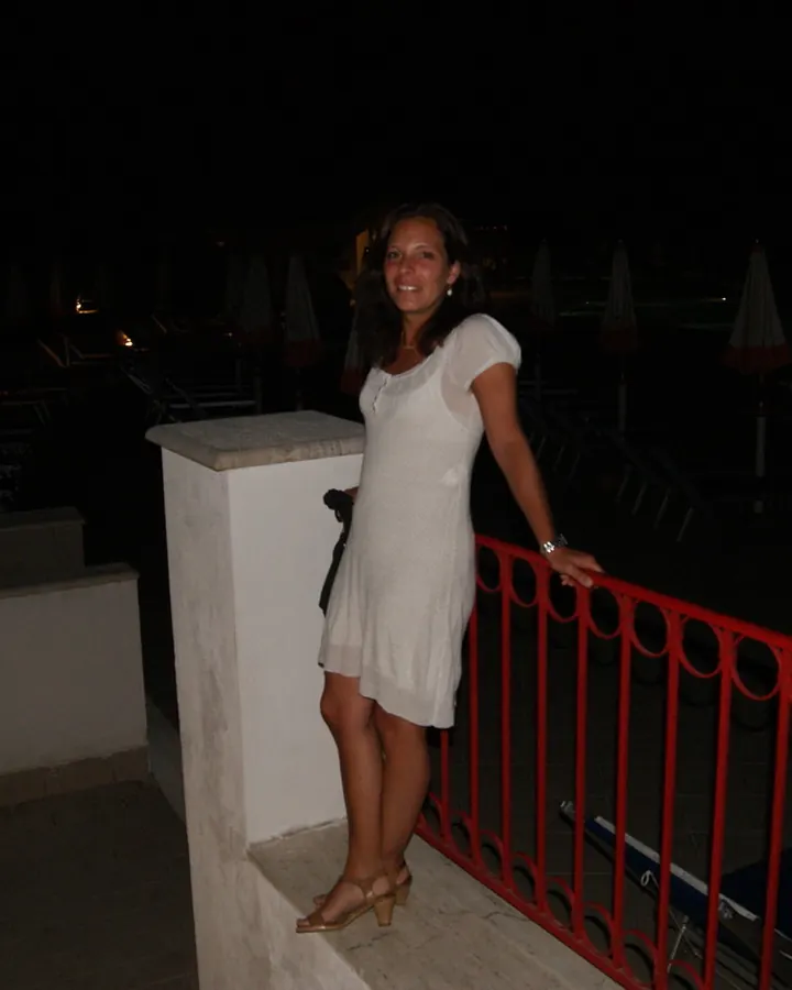 Femme en robe blanche debout sur un balcon la nuit, tenant une rambarde rouge, souriante.