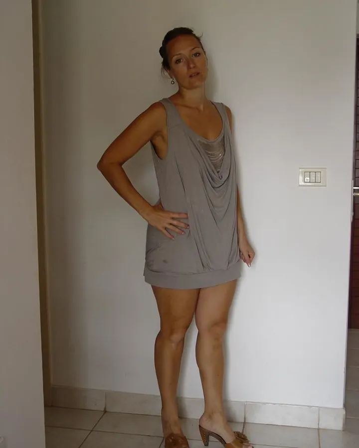Femme en robe grise, debout contre un mur, une main sur la hanche, regardant l'appareil.