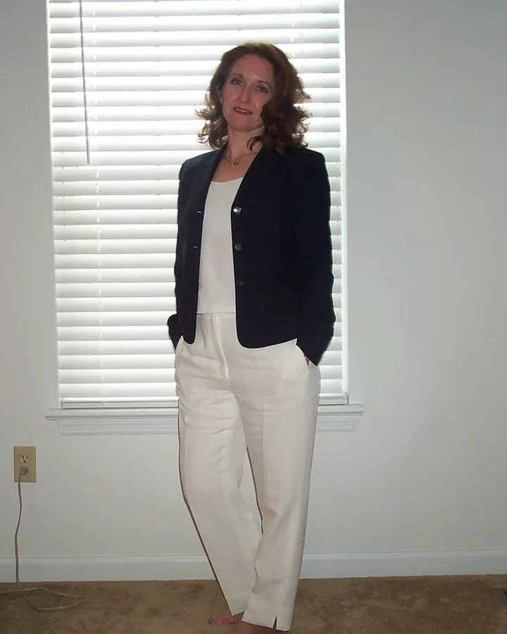 Femme en blazer noir et pantalon blanc, debout devant une fenêtre à persiennes