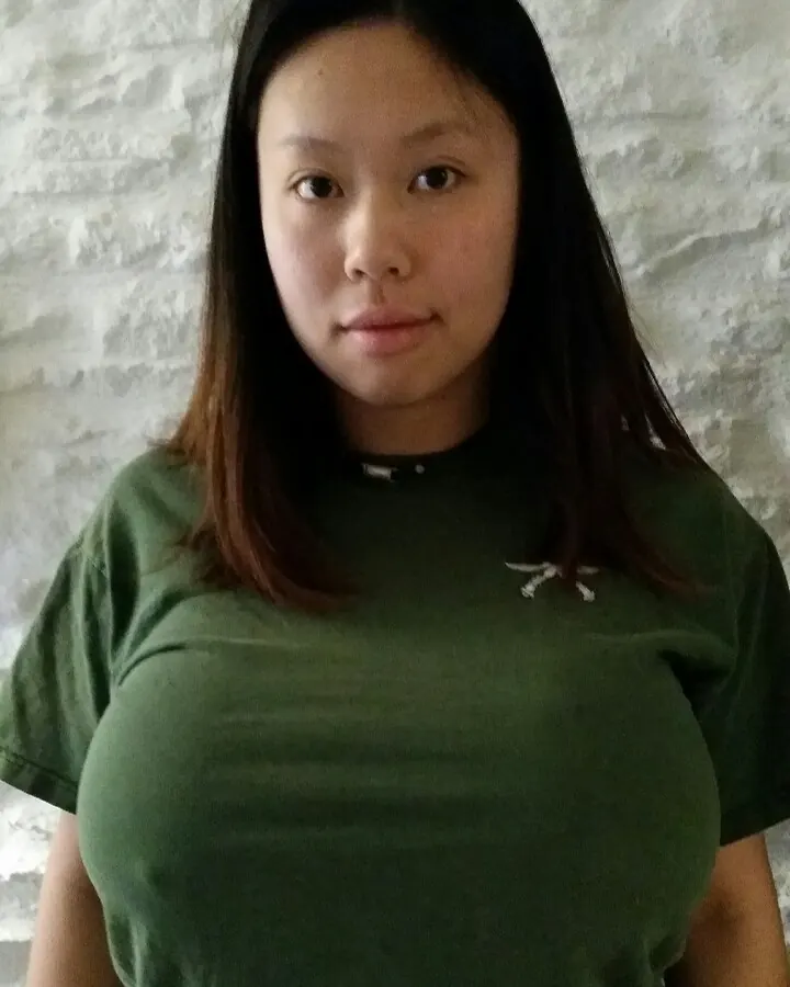 Femme asiatique en t-shirt vert, regard direct, poitrine marquée par le vêtement, devant un mur blanc.