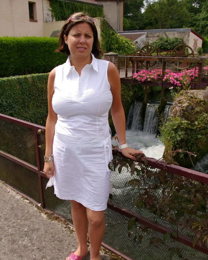 Femme aux cheveux châtains en robe blanche, debout près d'un petit pont, souriant, mains sur les hanches.