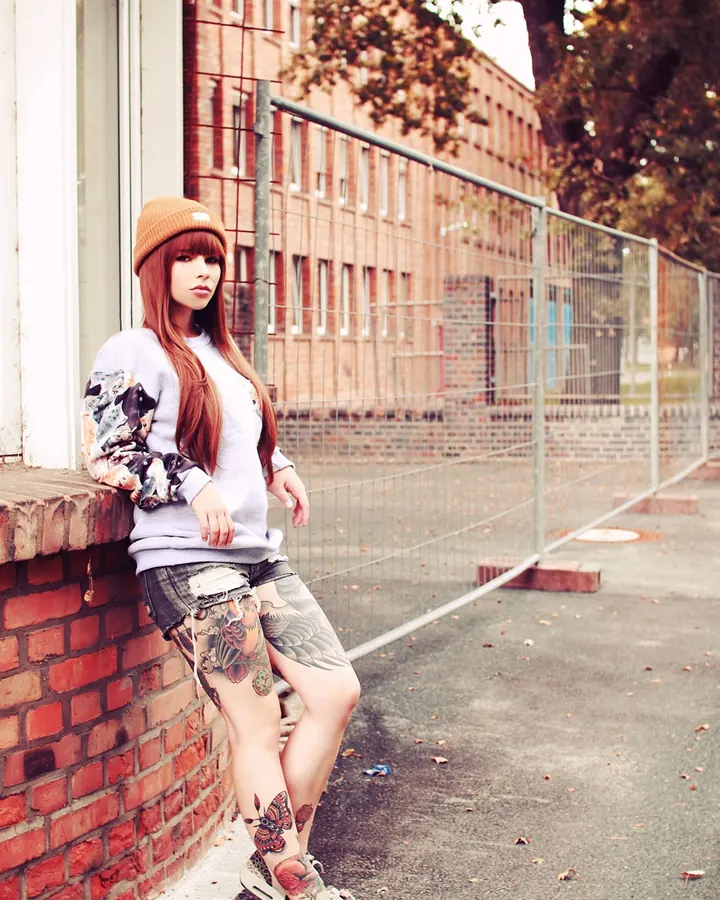 Femme asiatique aux cheveux roux, en pull gris, assise contre un mur, regard pensif, tatouages visibles.