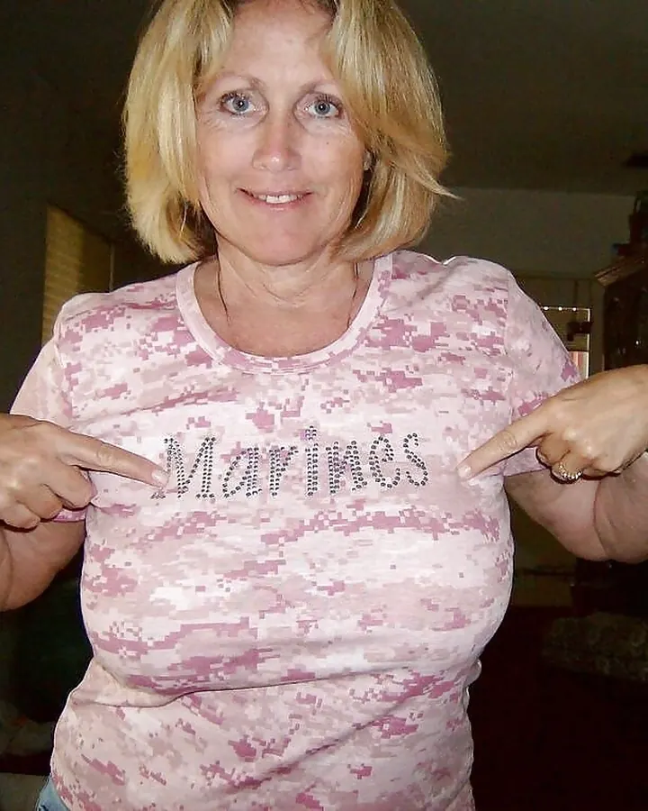 Femme blonde aux yeux bleus pointant vers le mot Marines brodé sur son t-shirt rose camouflage.